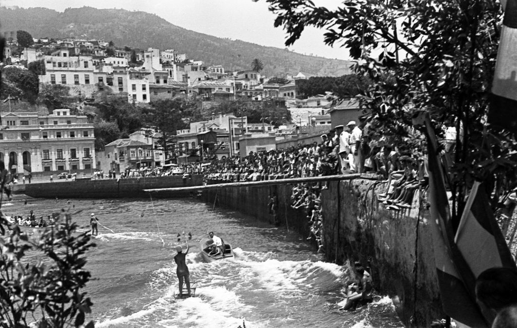 El malecón lleno de gente y el agua salpicada de bañistas, con la ciudad en la ladera al fondo